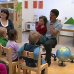 En far med et barn på fanget sitter foran en gruppe barn og gjør bevegelser til sang.