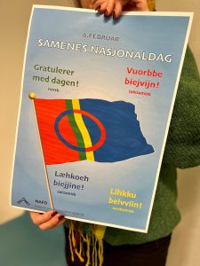 En plakat med overskriften 6. februar, Samenes nasjonaldag. Midt på plakaten er det et bilde av det samiska flagget, og rundt flagget står det Gratulerer med dagen på norsk og tre samiske språk.