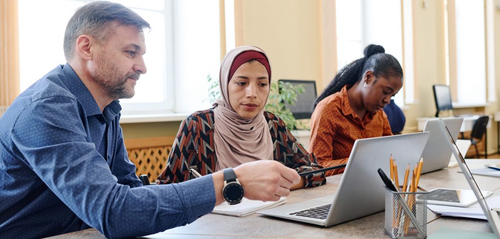 Mannlig lærer holder en blyant og peker på en laptop. Han hjelper en kvinnelig deltaker med hijab i klasserommet. Ved siden av sitter en annen kvinnelig deltaker foran en laptop.