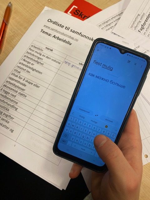 Foto av en ordliste med norsk ord. Ved siden av er det plass til å oversette ordene. Det er en mobil med ukrainsk oversettelse på.