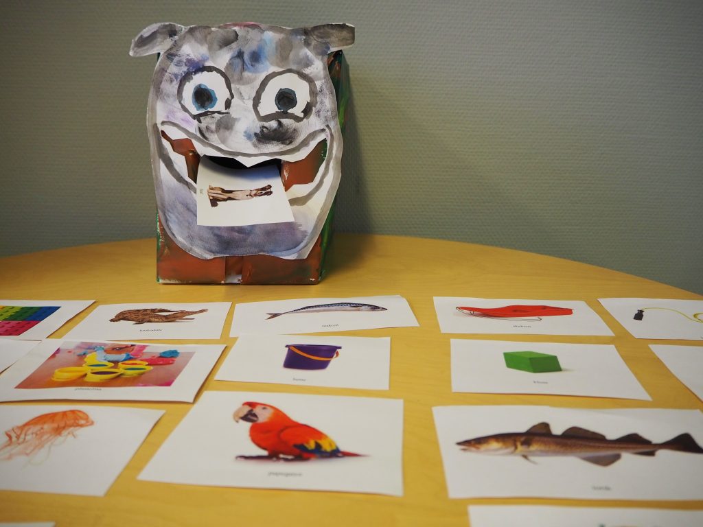 Foto av et bord med bildekort, kort med foto av ulike gjenstander. Bak bildekortene er et malt monsteransiktet som spiser et av bildekortene.