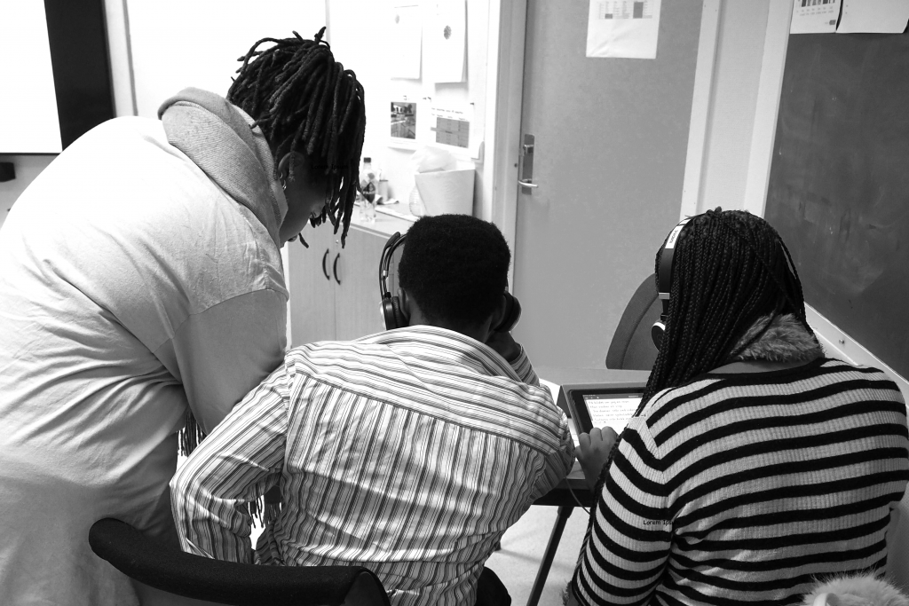 Svart-hvit foto av en flerspråklig assistent som står ved siden av to deltakere. Deltakerne deler bord og arbeider på nettbrett.