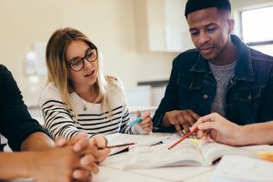 Foto av voksne mennesker sitter rundt et bord med bøker og skrivesaker foran seg. 
