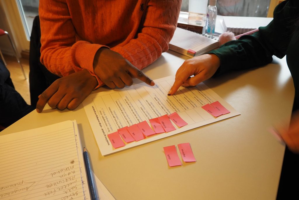 Foto av to deltakere som sitter ved et bord. På bordet ligger et ark med setninger i en tabell. Elevene legger rosa lapper med ord foran hver setning.