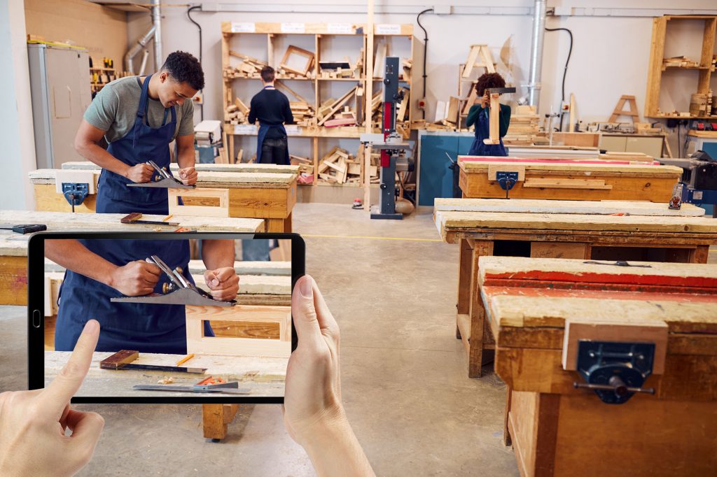 Foto av en sløydsal eller verksted. Flere elever står ved høvelbenker og jobber. En elev høvler. I forgrunnen holdes en ipad opp av to hender. På Ipaden er det foto av eleven som høvler.