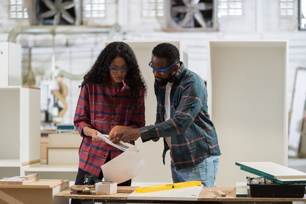 To elever står med en arbeidsbeskrivelse på et verksted. De ser på teksten og diskuterer. Foran dem er en høvelbenk med materiale og verktøy.