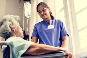 En eldre dame sitter i en rullestol. Ved siden av står en ung kvinne. Kvinna er praksiskandidat i helsearbeiderfaget.