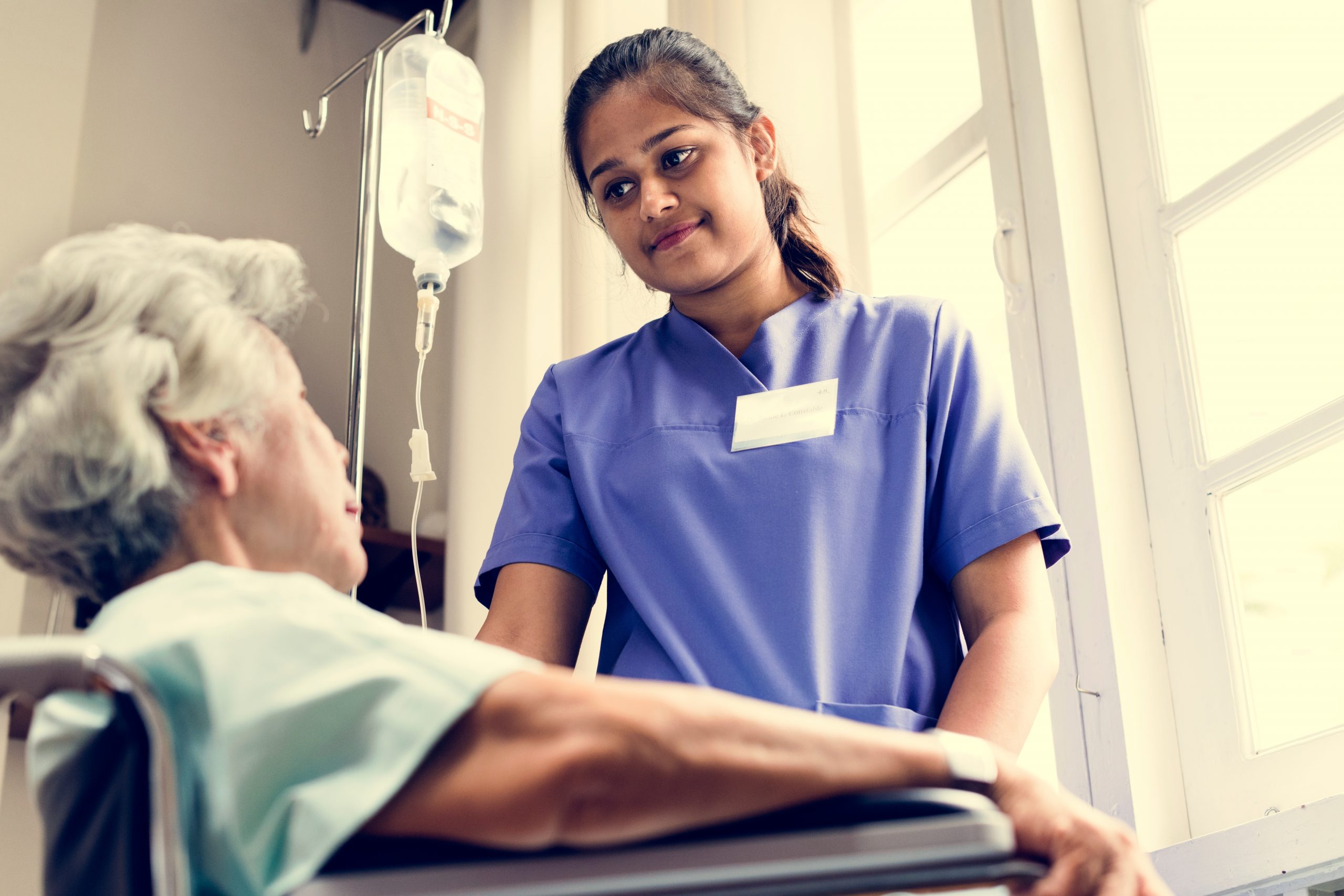 En eldre dame sitter i en rullestol. Ved siden av står en ung kvinne. Kvinna er praksiskandidat i helsearbeiderfaget.