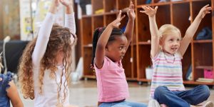 Foto av tre barnehagebarn som sitter på gulvet. Alle tre strekker armene i været som om det er bevegelser til en sang, vers eller lignende.