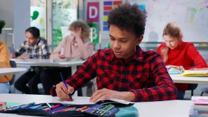 African-American teen student write in notebook at lesson in classroom. Schoolboy study sitting at desk in school
