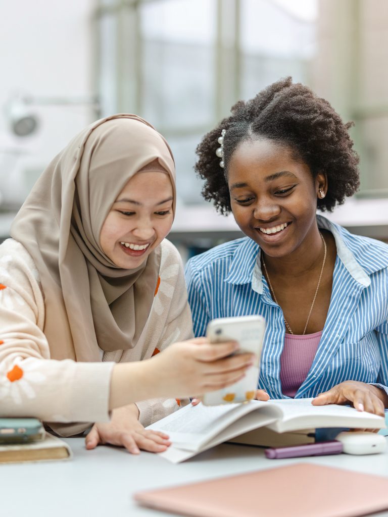 Foto av to elever i et klasserom. Foran seg har de en oppslått bok. Den ene holder opp en mobil. Bildet illustrerer at elevene kan hjelpe hverandre å oversette når de har et felles språk som ikke er norsk.