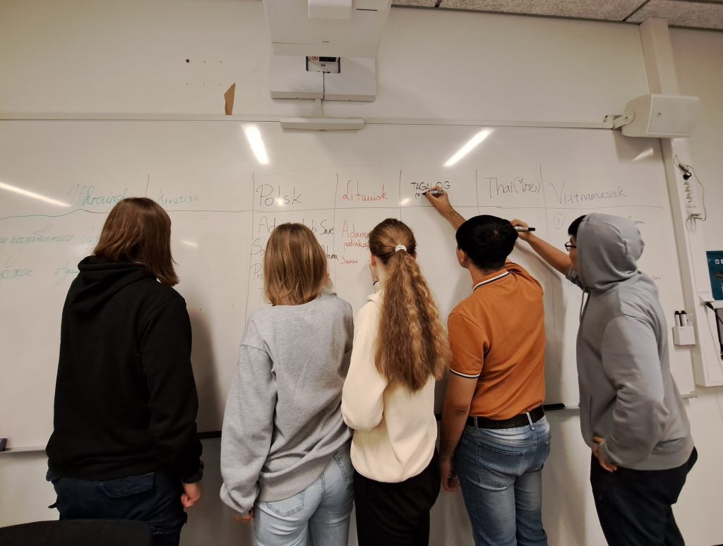 Foto av fem elever som står med ryggen til og skriver ord på flere språk på en tavle.