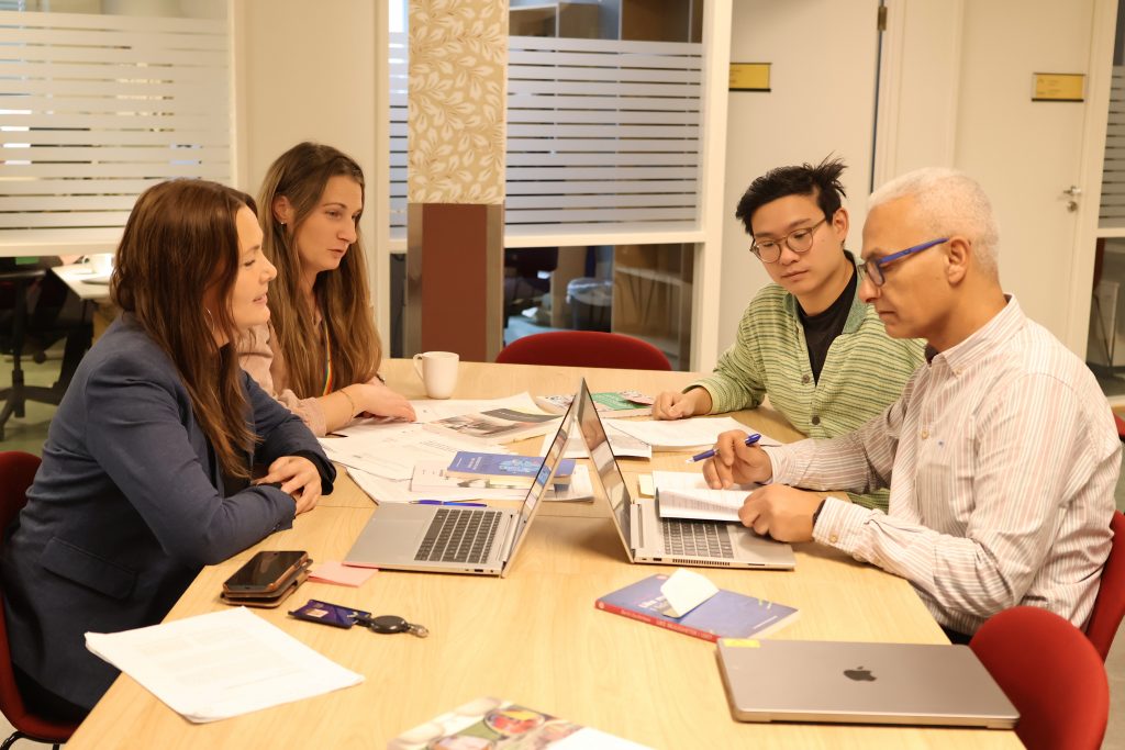 Fotografi av fire personer som sitter rundt et bord og jobber. På bordet ligger det papirer og bærbare datamaskiner.