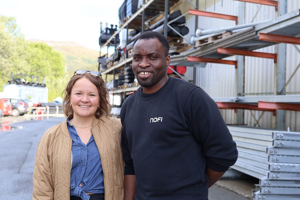 Foto av Kari Thomassen og Tuza Olukan. De står utenfor bedriften NOFI. I bakgrunnen ligger hyller med stillas og annet utstyr.