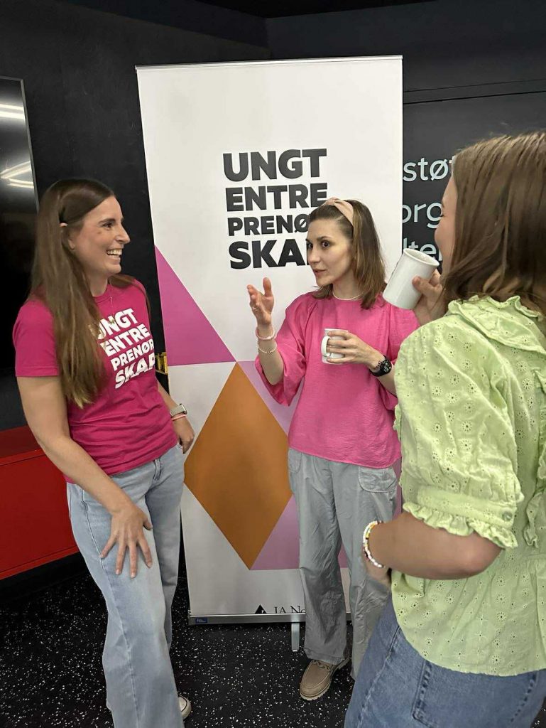 Fotografi av tre personer som smiler og snakker sammen foran en plakat / roll up med påskriften "Ungt entreprenørskap"