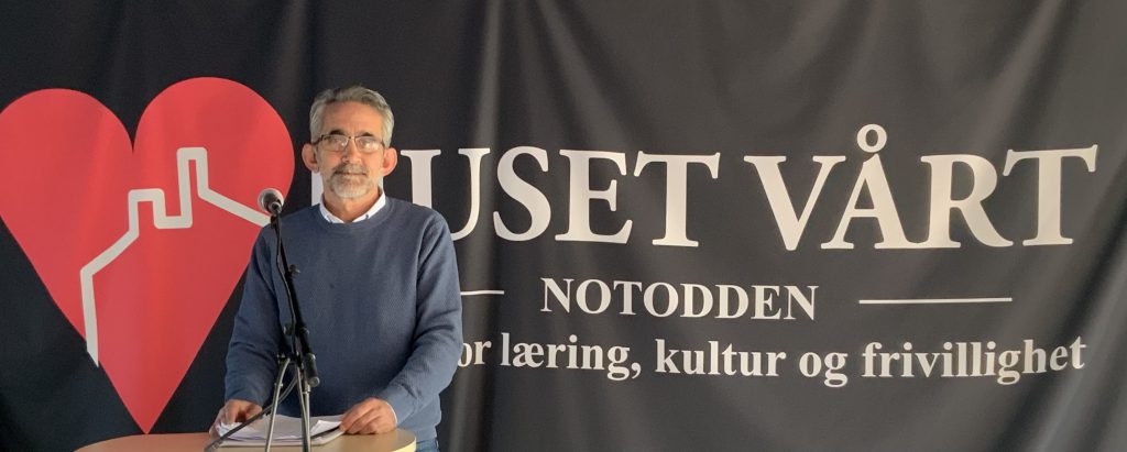 Fotografi av Ali Albakar som står på en talerstol. Bak ham henger det et banner med et stort rødt hjerte og skriften "Huset vårt".